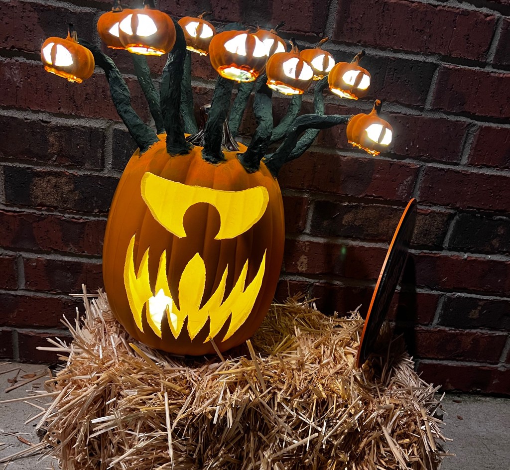 Pictured: my "behold o lantern." A foam craft pumpkin carved into a jack-o-lantern styled after the Dungeons and Dragons monster "the Beholder." It sits on a small hay bail in front of a brick wall. A single, narrowed eye has been carved into it, and a long-toothed, sinister, jagged grin. Green paper mache stalks extend from the top of the jack-o-lantern where smaller craft pumpkins each carved with a single cat-like eye. Each eye and the main beholder face are illuminated from within.