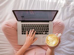 A photo from above of a white woman in pink pajama pants. She has a silver laptop open in her lap with her left hand on the keyboard. Her right hand holds a white cup of coffee with a leafy plant design drawn into the foam on the surface.