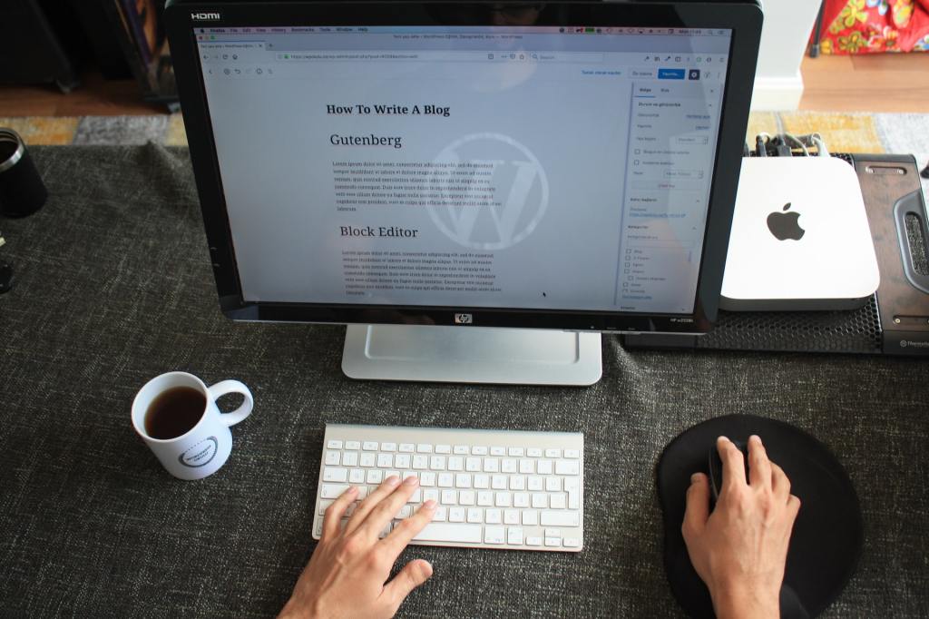 A top-down photo of an office computer desk with a mac mini seated on a gray linen-looking desk cover. Hands type on a tiny mac keyboard with a mostly full cup of black coffee off to the side. On the HP monitor, the WordPress blog post editor displays a sample blog post titled "How To Write A Blog" with large section headers reading "Gutenberg" and "Block Editor."