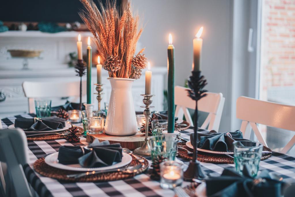A photo of a fancily dressed dining room table. White, farmhouse style chairs frame a table draped in a dark-green and white checkered tablecloth. Dark green napkins folded in a round, fanned shape sit atop white plates which sit atop wicker place mats. A bundle of wheat and pine cones in a white vase on a wooden slab centerpiece is flanked by white candles in tall, silver candleholders, which are flanked by dark green candles in white candleholders, which are flanked by short white candles sitting in pine cone shaped candleholders.